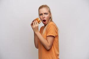 Shocked young attractive long haired blonde woman with ponytail hairstyle holding big burger in her hands and looking discomposedly at camera, frowning eyebrows with opened mouth over white background photo