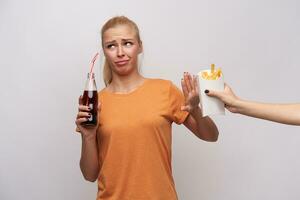 Displeased young blonde female with casual hairstyle looking aside with pout and frowning her face with raised palm, drinking soda and refusing to eat french fries, isolated over white background photo