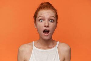 Close-up of surprised young female wearing her red hair in knot over orange background, wearing white top, looking to camera with wide mouth opened and rounding eyes amazedly photo