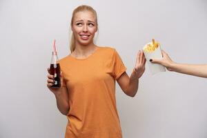 Confused young pretty blonde lady with ponytail hairstyle frowning eyebrows and raising hand with refusing gesture while someone is offering her french fries, isolated over white background photo