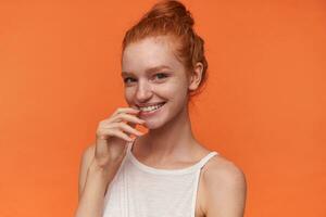 Portrait of positive young redhead woman with knot wearing white top, looking at camera cheerfully and touching underlip with index finger, posing over orange background photo