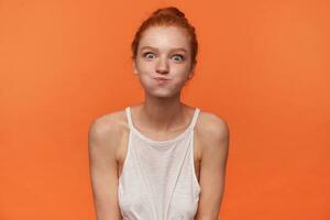 Indoor photo of young funny lady wearing her red hair in knot, looking at camera and puffing out cheeks, fooling and making faces over orange background in white top