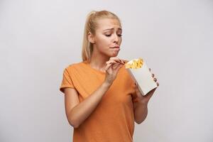 Indoor shot of young long haired blonde lady keeping paper box with french fries and looking excitedly at it, wanting to eat it, but worrying about extra calories, isolated over white background photo