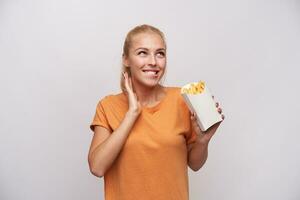 Portrait of happy young lovely blonde woman with casual hairstyle biting her underlip and foretasting like she is eating french fries, smiling cheerfully over white background photo