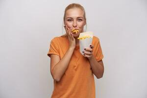 Shot of positive young pretty blonde female with casual hairstyle looking cheerfully at camera with mouth full of french fries, being hungry and happy to get meal, isolated over white background photo