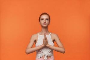 interior Disparo de bonito joven hembra con astuto bollo peinado vistiendo casual ropa, mirando hacia arriba con calma rostro, acuerdo manos doblada en namaste gesto, posando terminado naranja antecedentes foto