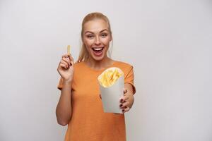 Studio photo of happy attractive young blonde female rejoicing about fresh french fries in her hand and looking joyfully at camera with broad smile, posing against white background