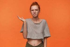 Studio photo of pretty young female wearing her foxy hair in knot, frowning face with pout and raising palm up, standing over orange background in grey t-shirt and green shorts