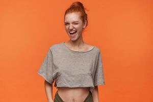 Portrait of overjoyed young readhead woman wearing hair in knot posing over orange background, smiling cheerfully to camera and winking with one eyes, wearing casual clothes photo