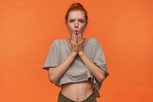 Horizontal shot of shocked young readhead lady with hair in knot standing over orange background in casual clothes, rounding lips and holding hands on face photo