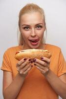 Close-up of hungry blue-eyed excited young blonde lady with tasty hot dog in her hands looking cheerfully at camera and licking her lips, standing against white background photo