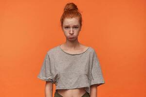 Portrait of cute offended young redhead woman with bun hairstyle wearing casual clothes, posing over orange background with hands down, wrinkling forehead with pursed lips photo