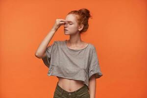 Studio photo of attractive tired young woman in casual clothes holding her nose bridge with hand, posing over orange background with closed eyes, wearing her red hair in knot