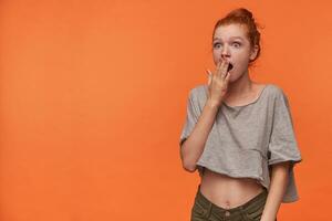 Image of shocked young readhead lady with foxy hair in knot wearing grey t-shirt and green shorts, covering mouth amazedly with raised palm, isolated over orange background photo