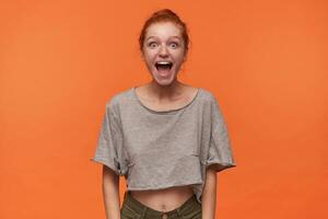 alegre joven cabeza lectora mujer con bollo peinado vistiendo gris camiseta y verde pantalones cortos , en pie terminado naranja antecedentes con manos abajo. positivo humano emoción facial expresión cuerpo idioma foto