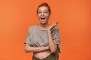 Studio photo of lovely joyful young readhead woman with bun hairstyle wearing grey t-shirt and green shorts, looking surprisedly to camera with wide mouth opened, standing over orange background