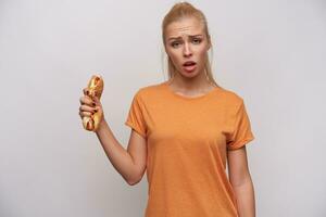 Naughty young long haired blonde woman with ponytail hairstyle looking defiantly at camera and frowning eyebrows, holding smashed hot dog in raised hand while standing over white background photo
