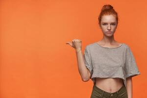 Indoor shot of charming young readhead lady in grey t-shirt and green shorts posing over orange background, wearing her foxy hair in knot, raising thumb and pointing aside photo