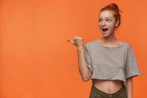 Joyful young readhead lady with bun hairstyle looking surprisedly aside, raising thumb and pointing away with wide cheerful smile, isolated over orange background in grey t-shirt and green shorts photo