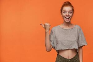 Overjoyed young redhead female in grey t-shirt and green shorts posing over orange background, showing with raised thumb aside, smiling broadly and demonstrating her white perfect teeth photo