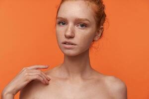 Indoor shot of attractive young female wearing her red hair in bun hairstyle standing over orange background, looking at camera gently with ajar mouth and touching shoulder with raised hand photo