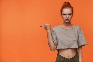 Portrait of serious young readhead woman lwearing her hair in bun, frowning scowlingly to camera and raising hand with thumb pointing aside, standing over orange background photo