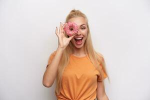 Indoor shot of joyful attractive young blonde woman with casual hairstyle making fun with donut in raised hand while looking cheerfully at camera, isolated over white background photo