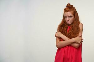 Indoor shot of frightened young readhead female with wavy hair wearing no make up, hugging herself with arms and looking aside with empty eyes, isolated over white background photo