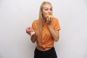 Excited young pretty long haired blonde woman looking poisitively at camera with raised eyebrows and tasting donats in her hands, wearing casual clothes over white background photo