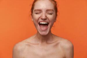 retrato de joven lleno de alegría atractivo dama vistiendo su rojo pelo en bollo posando terminado naranja antecedentes con cerrado ojos, demostración su agradable emociones con amplio alegre sonrisa foto