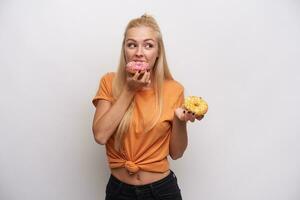 Positive slim young blonde female with casual hairstyle holding donats and biting piece while looking aside, dressed in orange t-shirt while posing against white background photo