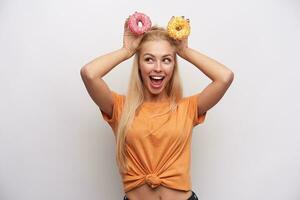 gracioso retrato de joven hermosa rubia mujer en naranja camiseta haciendo ratón orejas desde dona en elevado manos y sonriente felizmente mientras engañando terminado blanco antecedentes foto