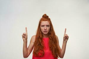 Photo of beautiful young woman with foxy wavy hair wearing no make up posing over white background, looking to camera with confused face and raising forefingers, frowning with wrinkled forehead