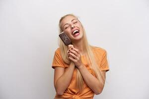 Studio photo of young good looking long haired blonde lady in orange t-shirt smiling happily with closed eyes and fooling with ice-cream in her hands, isolated over white background
