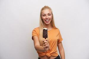 interior Disparo de joven encantador alegre largo peludo rubia dama con helado en palo sonriente felizmente a cámara, siendo en bonito estado animico mientras posando en contra blanco antecedentes foto