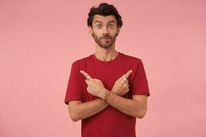 Surprised young man with trendy haircut pointing with forefingers in different directions, standing over pink background in casual clothes, looking at camera with raised eyebrows and biting underlip photo