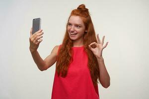 Portrait of charming young lady in pink dress wearing her foxy hair in bun, making photo of herselg with mobile phone, smiling widely to camera and rasing hand with ok gesture