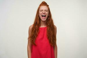 Indoor photo of pretty excited young lady with foxy bun hairstyle wearing pink dress, posing over white background with hands down, shouting loud with closed eyes