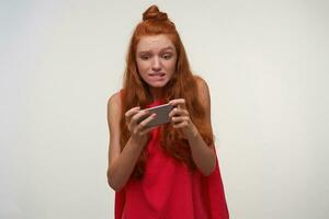 Photo of young agitate woman wearing her foxy hair in bun, playing games on her smartphone, looking at screen with excited face and biting underlip, isolated over white background