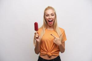 Overjoyed lovely young blonde woman with casual hairstyle pointing with forefinger on berry ice-cream in her hand, looking joyfully to camera with wide mouth opened, isolated over white background photo