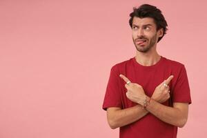 Portrait of attractive young man with trendy haircut pointing with index fingers in different directions, standing over pink background in red t-shirt, looking aside and showing tongue photo