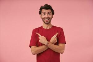 Overjoyed young pretty man with trendy haircut showing with index fingers in different directions, looking to camera happily with wide mouth opened and raising eyebrows, isolated over pink background photo