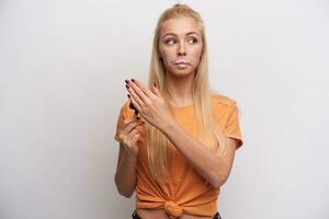 Indoor photo of pretty young blonde female wearing casual hairstyle while posing over white background, hiding ice-cream with soiled face, pretending that she did not eat it