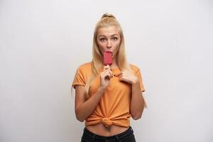 perplejo joven bonito rubia hembra con cola de caballo peinado comiendo helado y mirando confusamente a cámara, vestido en naranja camiseta mientras en pie terminado blanco antecedentes foto