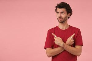 Indoor shot of handsome dark haired young male looking aside with doubting face, showing with index fingers in different sides while posing over pink background photo