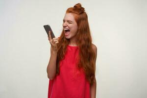 Stressed young pretty lady with foxy hair in bun hairstyle holding mobile phone in hand over white background, shouting with wide mouth opened to handset, wearing pink dress photo