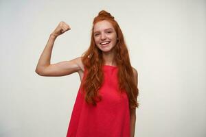 Studio photo of charming young readhead lady with bun hairstyle looking to camera with wide cheerful smile, raising hand and demonstrating her strong biceps, isolated over white background