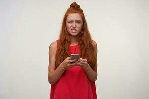 Portrait of unpleased young readhead lady pink dress wearing her hair in knot, standing over white background with mobile phone in hands, frowning and looking to camera with pout photo