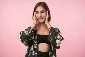 Positive young pretty brown-eyed brunette lady holding her face with raised hands and smiling pleasantly while looking at camera, isolated over pink background photo