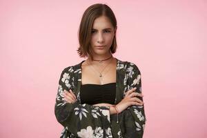 Serious young attractive brunette lady with bob haircut keeping her hands crossed while looking at camera with folded lips, standing over pink background photo